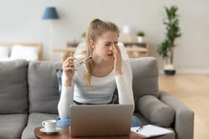 A woman rubbing her eyes and holding her glasses with a laptop in front of her